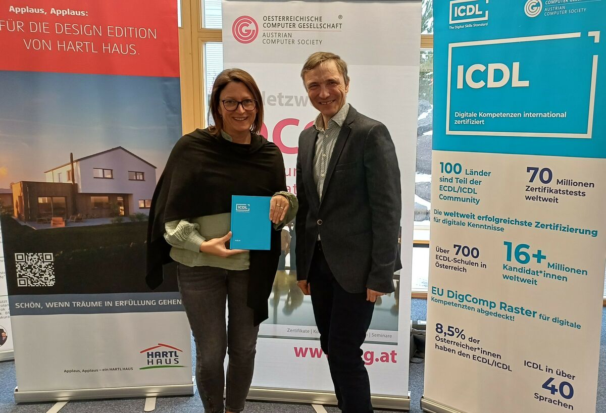 Sonja Früchtl und Ronald Bieber. Sonja Früchtl hält ein ICDL Notizbuch, im Hintergrund Roll-ups von Hartl Haus, ICDL und OCG