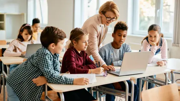 Lehrerin zeigt in einer hellen Schulklasse vier Schüler*innen etwas am Laptop