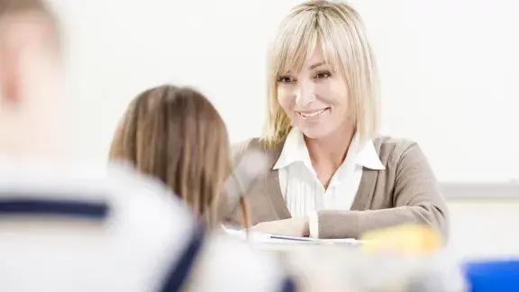 Frau schaut auf zwei sitzende Schüler*innen, deren Rückenansicht man nur verschwommen sieht.