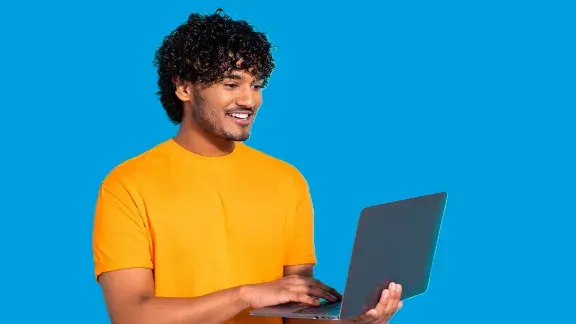 Junger Mann, dunkle Haare, dunkler Teint, gelbes T-Shirt mit aufgeklappten Laptop blickt gut gelaunt auf Bildschirm