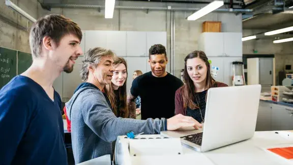 Team junger Mitarbeiter*innen steht um etwas älteren Mann, der etwas am Laptop erklärt, Raum sieht nach Industriehalle aus