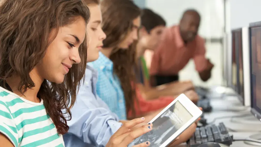 Mädchen am Tablet im Hintrgrund drei weitere Schüler*innen am Computer arbeitend sowie ein Lehrer, der gerade etwas zu erklären scheint