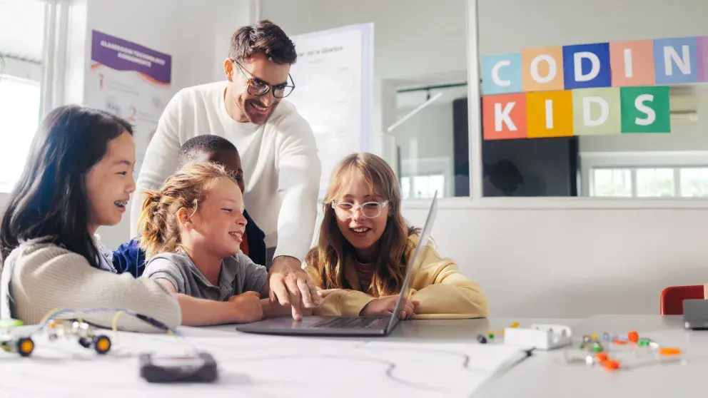 Drei Mädchen arbeiten auf einem Tablet, ein Junge im Hintergrund, freundlicher Lehrer erklärt etwas, helles Klassenzimmer, in bunten Buchstaben steht auf der Fensterscheibe: Coding Kids
