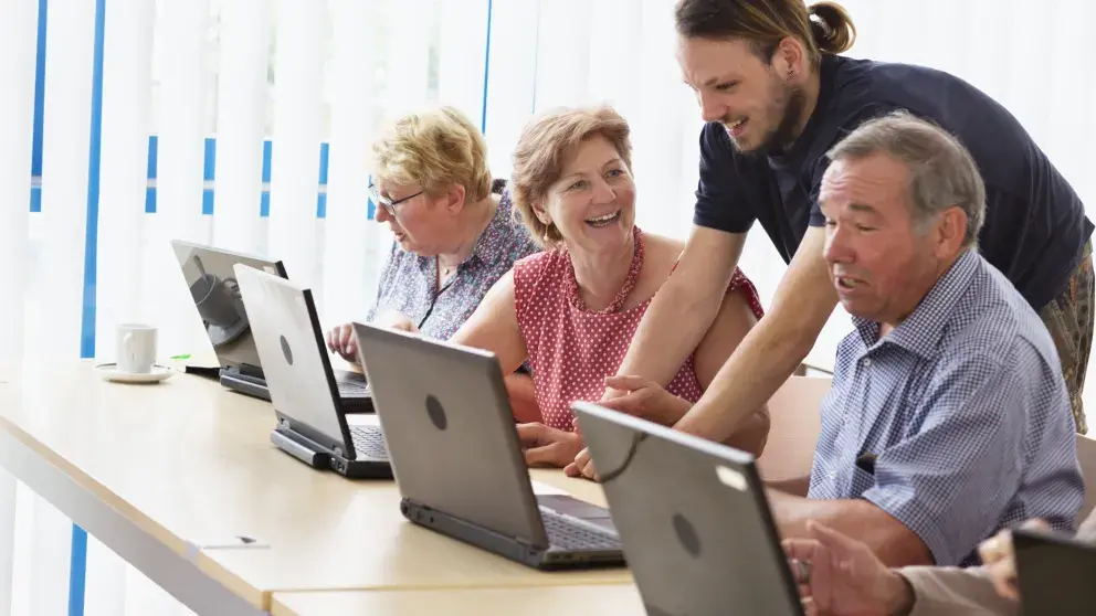 Junger Mann erklärt drei älteren Personen, die vor  Laptops sitzen etwas