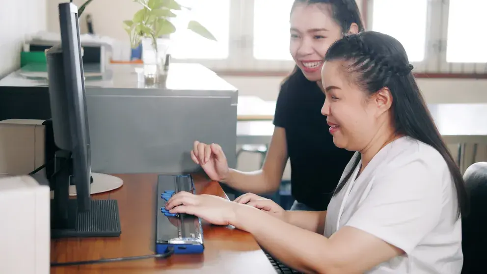 Glückliche asiatische Frauen am Arbeitsplatz im Büro, einschließlich einer blinden Person, die einen Computer mit einer aktualisierbaren Braillezeile als Hilfsmittel benutzt. Inklusion von Behinderten bei der Arbeit Konzepte.