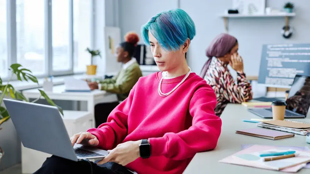 asiatisch aussehende Jugendliche Person mit blauen Haaren und Laptop, in einem Büro im Hintergrund zwei Kolleginnen mit dunkler Hautfarbe und eine mit Kopftuch, Diversität