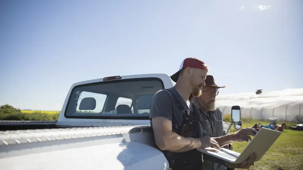 Zwei Bauern lehnen an einem Pick-up-Auto auf einem Feld und nutzen Laptop und ein weiteres technisches Gerät