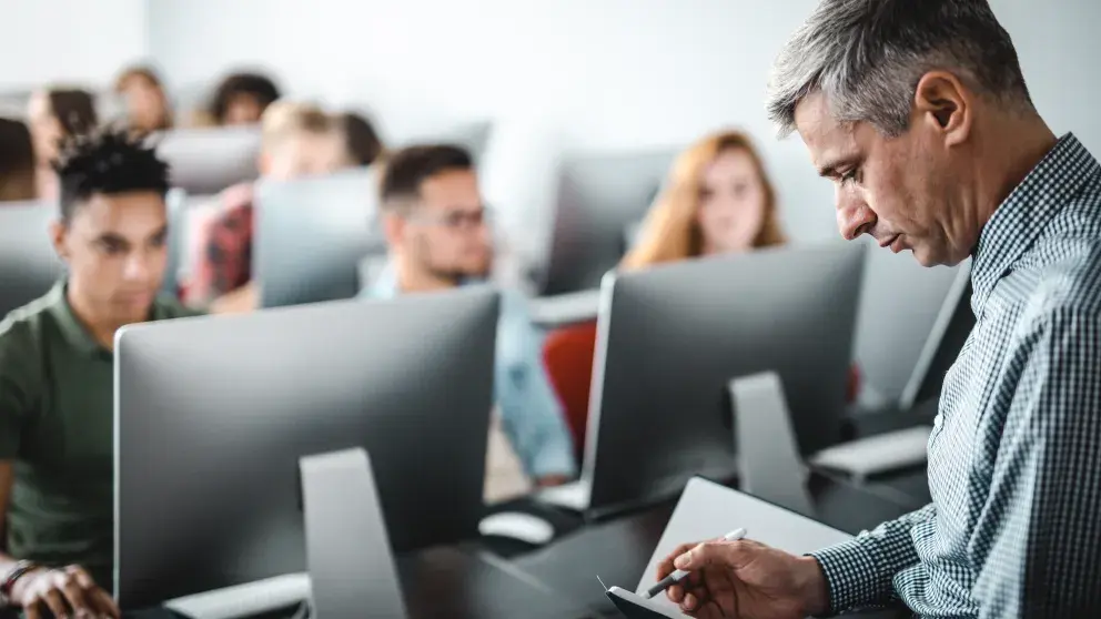 Test Center Trainer (grauhaariger Mann) macht sich Notizen vor einer Klasse, die vor Bildschirmen sitzt