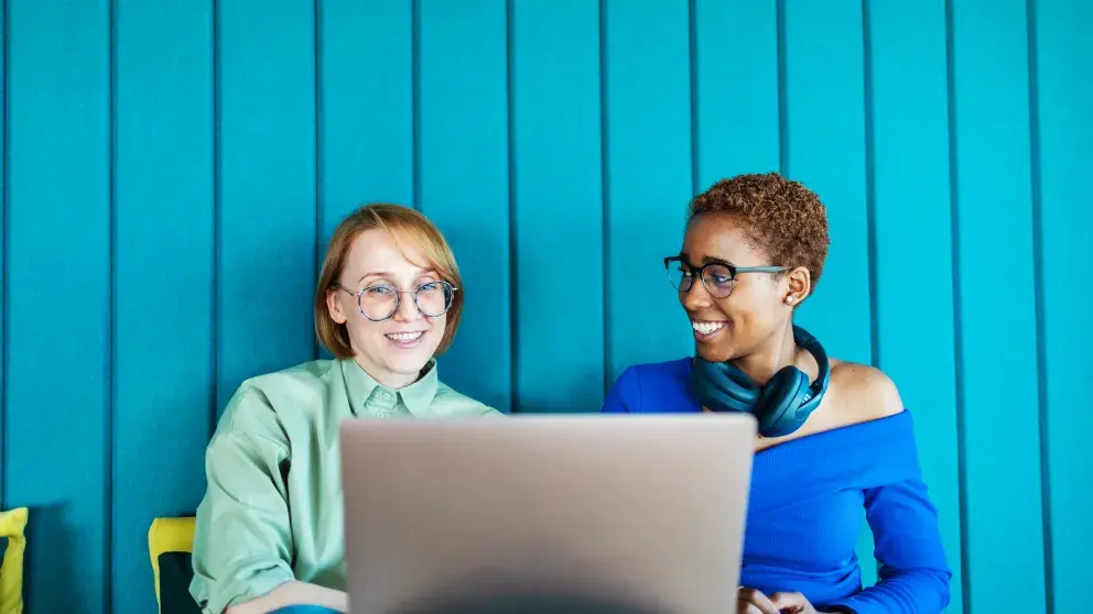 Zwei junge Frauen vor einem Laptopbildschirm gut gelaunt, unterhalten sich